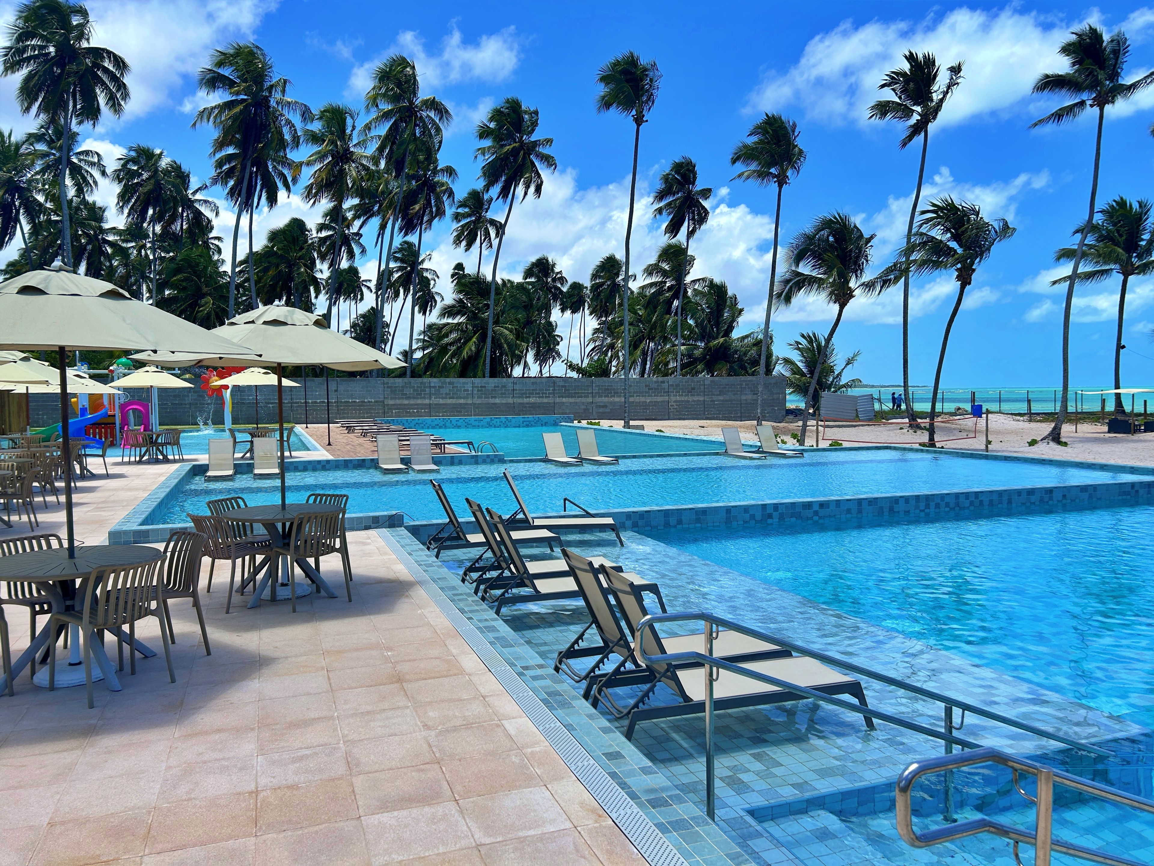 A pool area with lounge chairs and umbrellas, surrounded by palm trees and a clear sky.