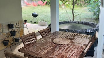 Un espacio de comedor al aire libre techado, con una mesa y sillas de madera, una parrilla y vistas a un jardín.