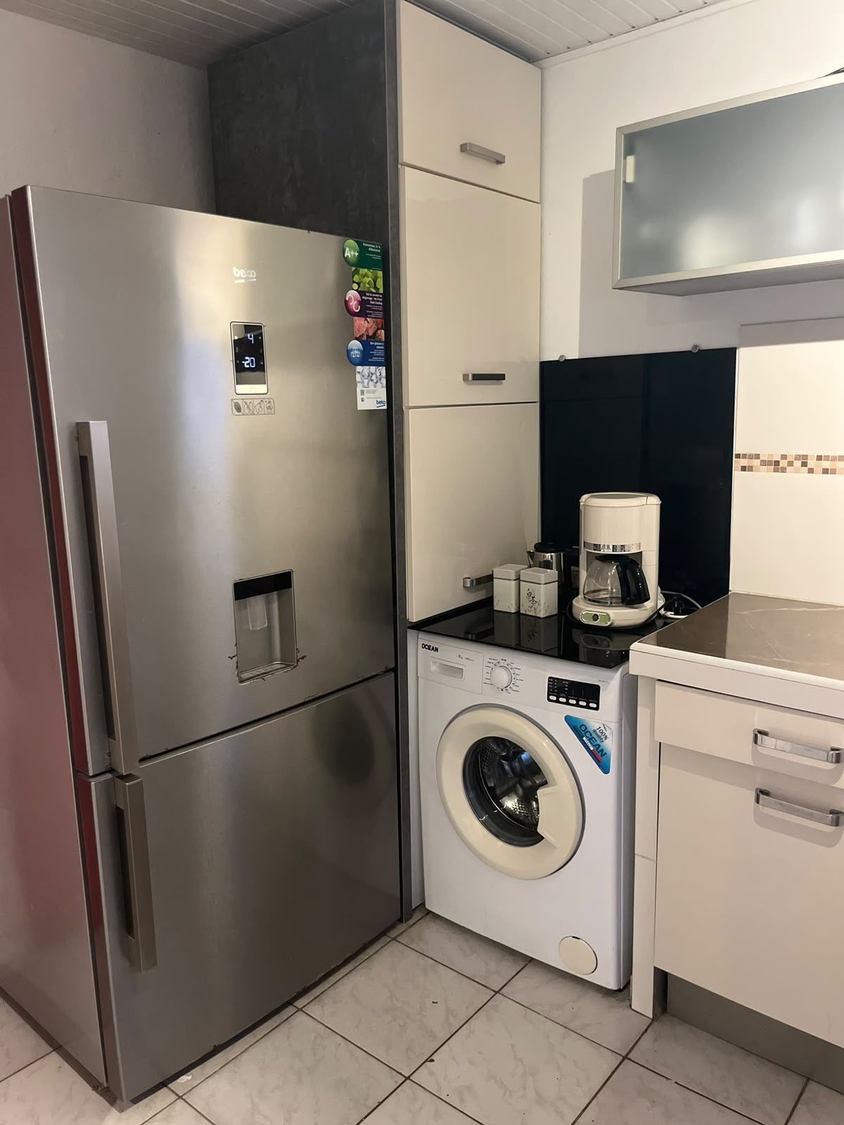 Eine kleine Küche mit einem Edelstahlkühlschrank, einer Waschmaschine und einer Kaffeemaschine.