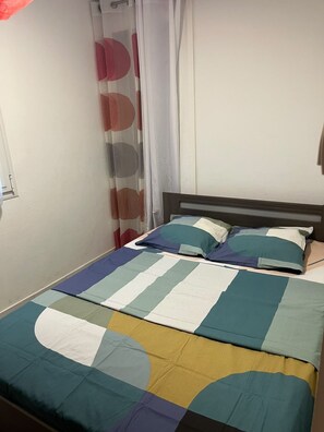 Una cama individual con un edredón colorido y dos almohadas, una ventana con cortina y una pared lisa.