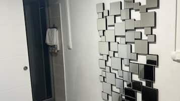 Un baño con piso de baldosas, ducha, espejo en la pared y cesto de ropa.