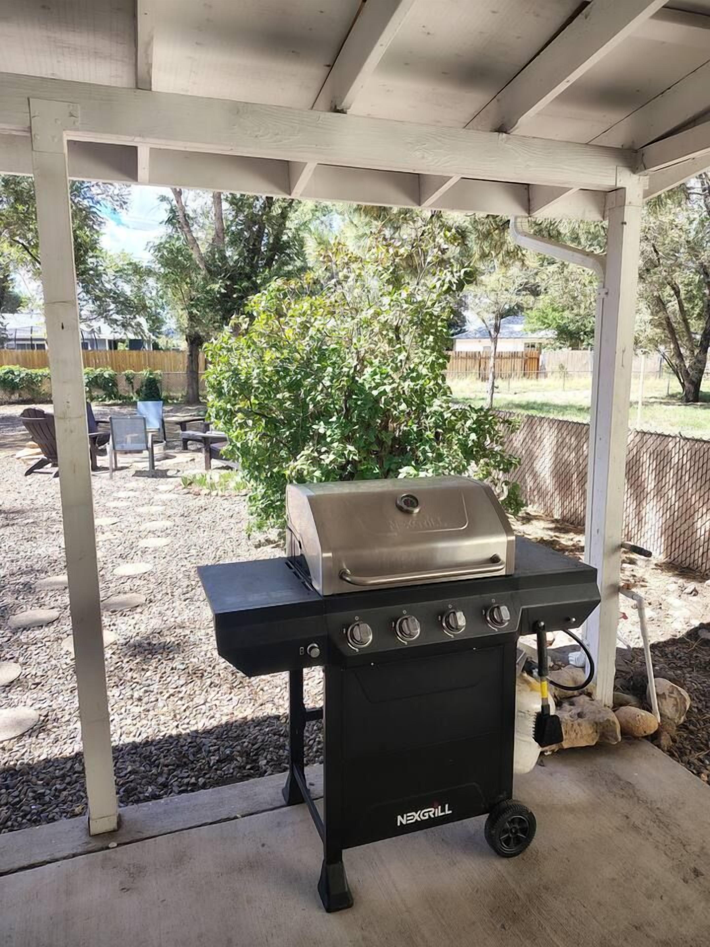 Una parrilla de barbacoa bajo un porche cubierto, con suelo de grava y follaje al fondo.