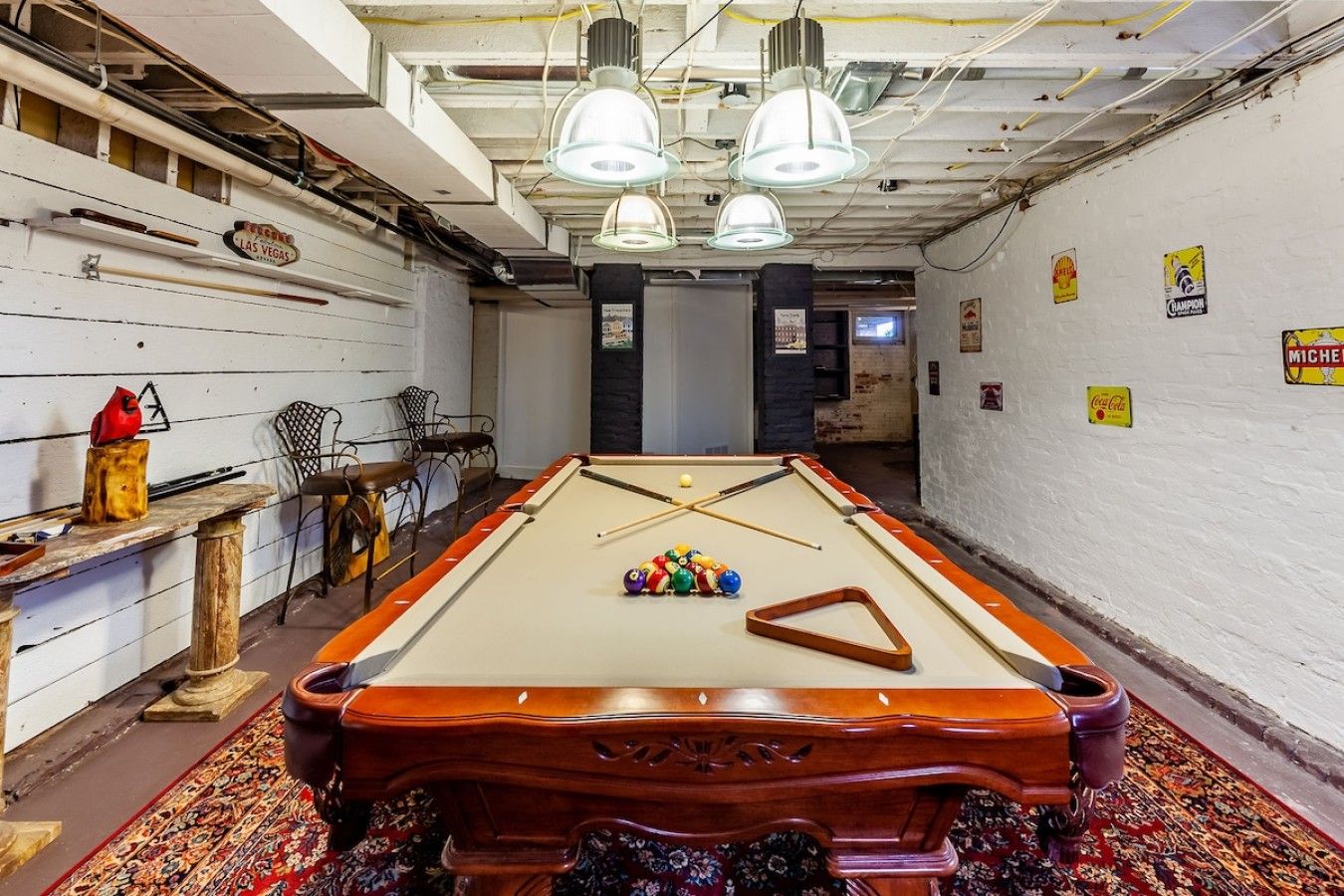 A pool table with billiard balls and cues, a wooden chair, and a table with a bottle and cue stick.