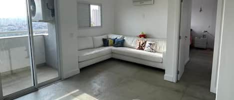 A white living room with a white sofa, a glass door leading outside, and a small table with a chair.