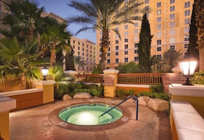 Zona de spa con un jacuzzi rodeado de palmeras y un edificio al fondo.