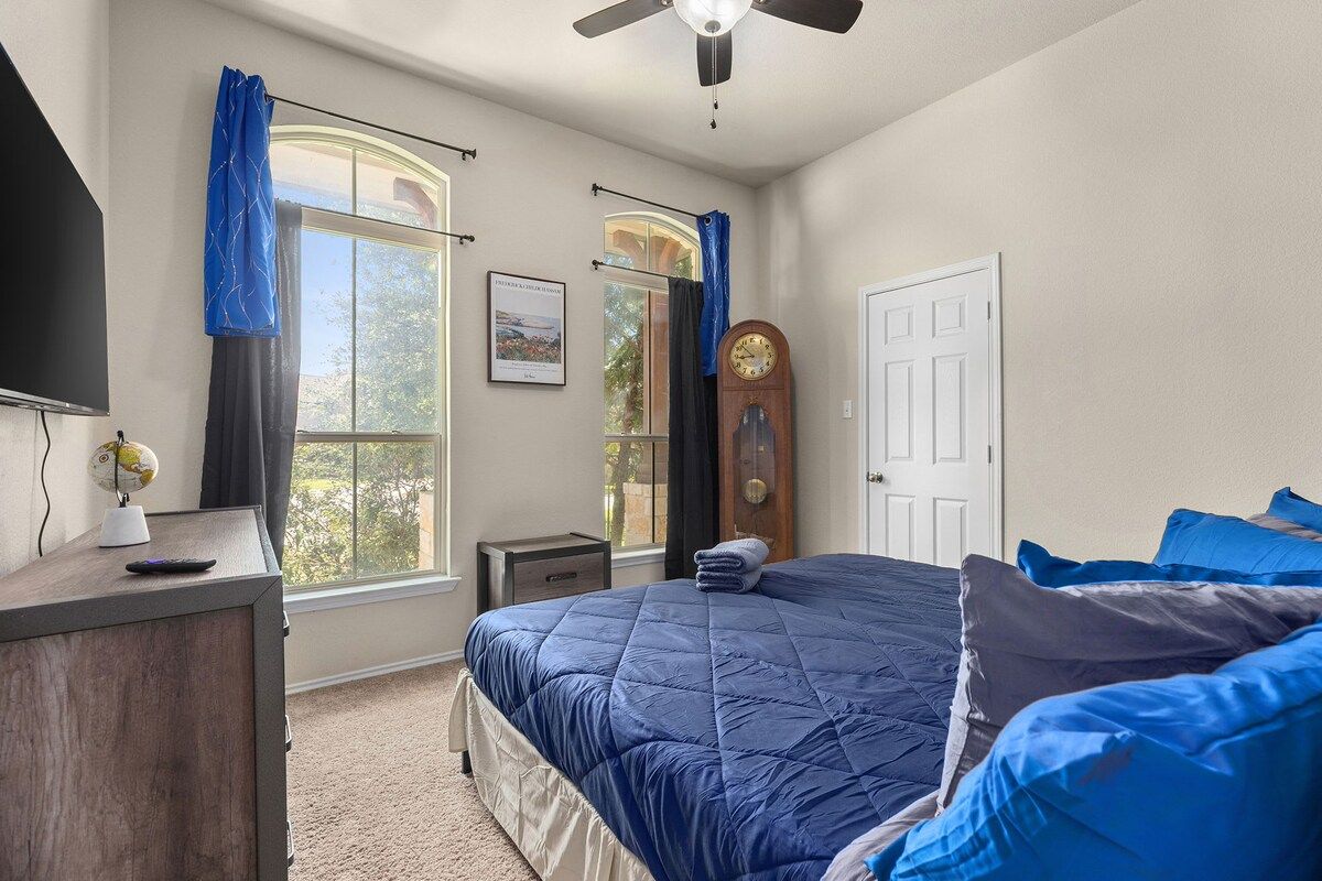 A bedroom with a bed, a desk, a television, a clock, and a window with curtains.