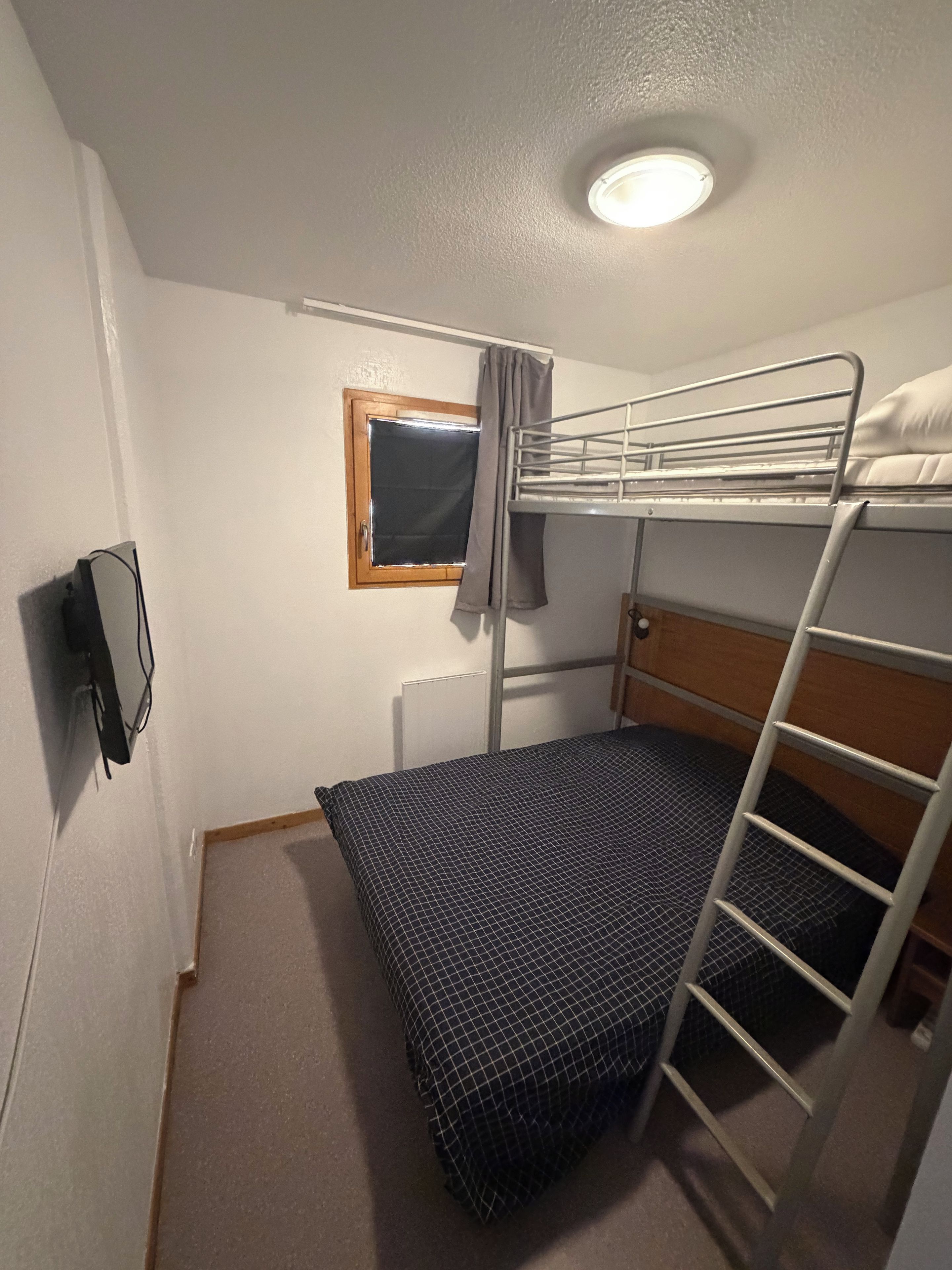 A small, single-bed room with a bunk bed, a window with a curtain, a wall-mounted TV, and a ceiling light.