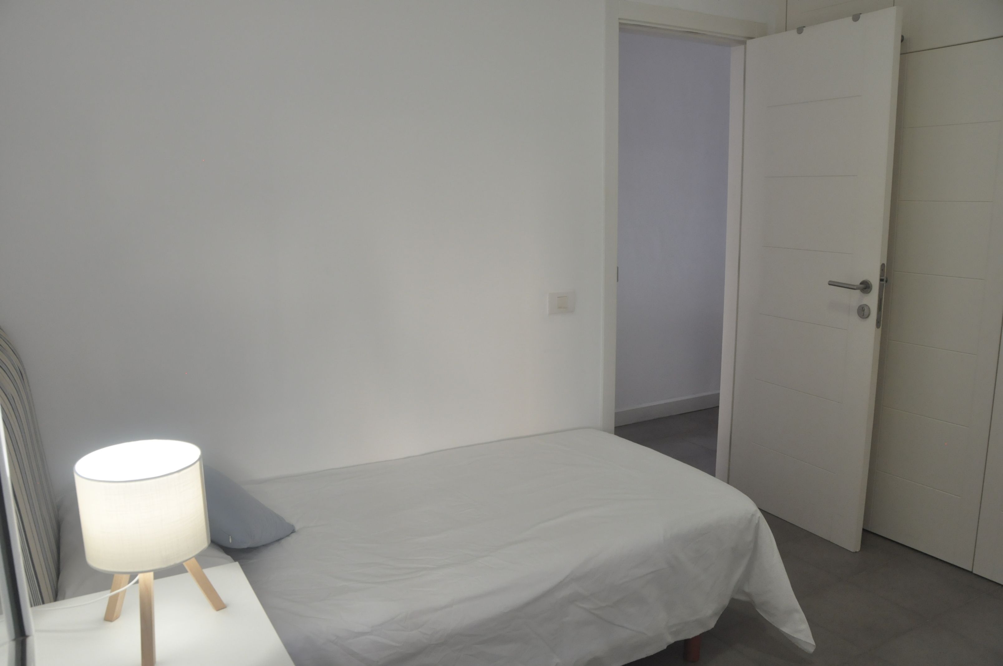 A bedroom with a single bed, a bedside table with a lamp, and a door leading to another room.