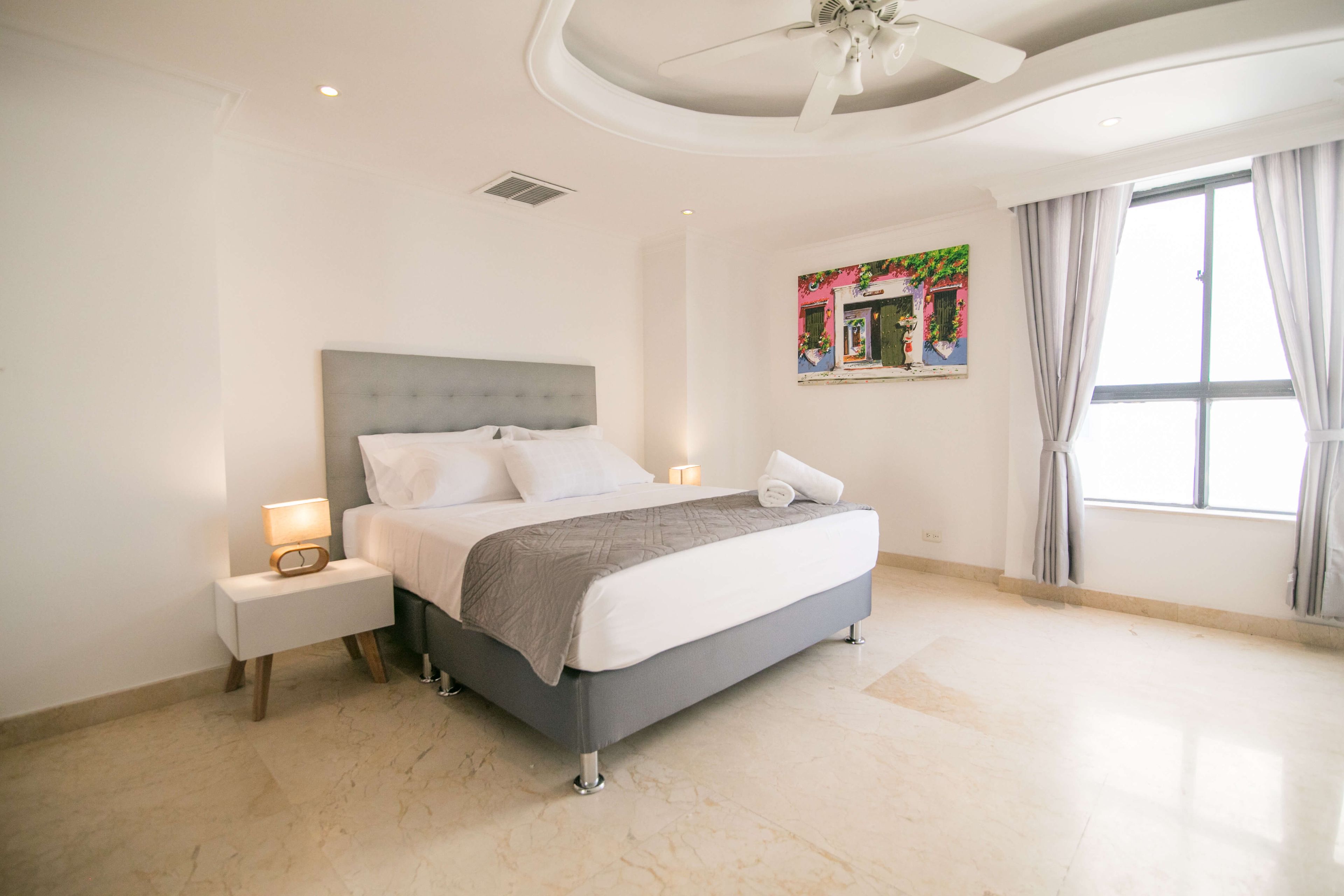 A hotel room with a bed, nightstands, a ceiling fan, a window with curtains, and a painting on the wall.