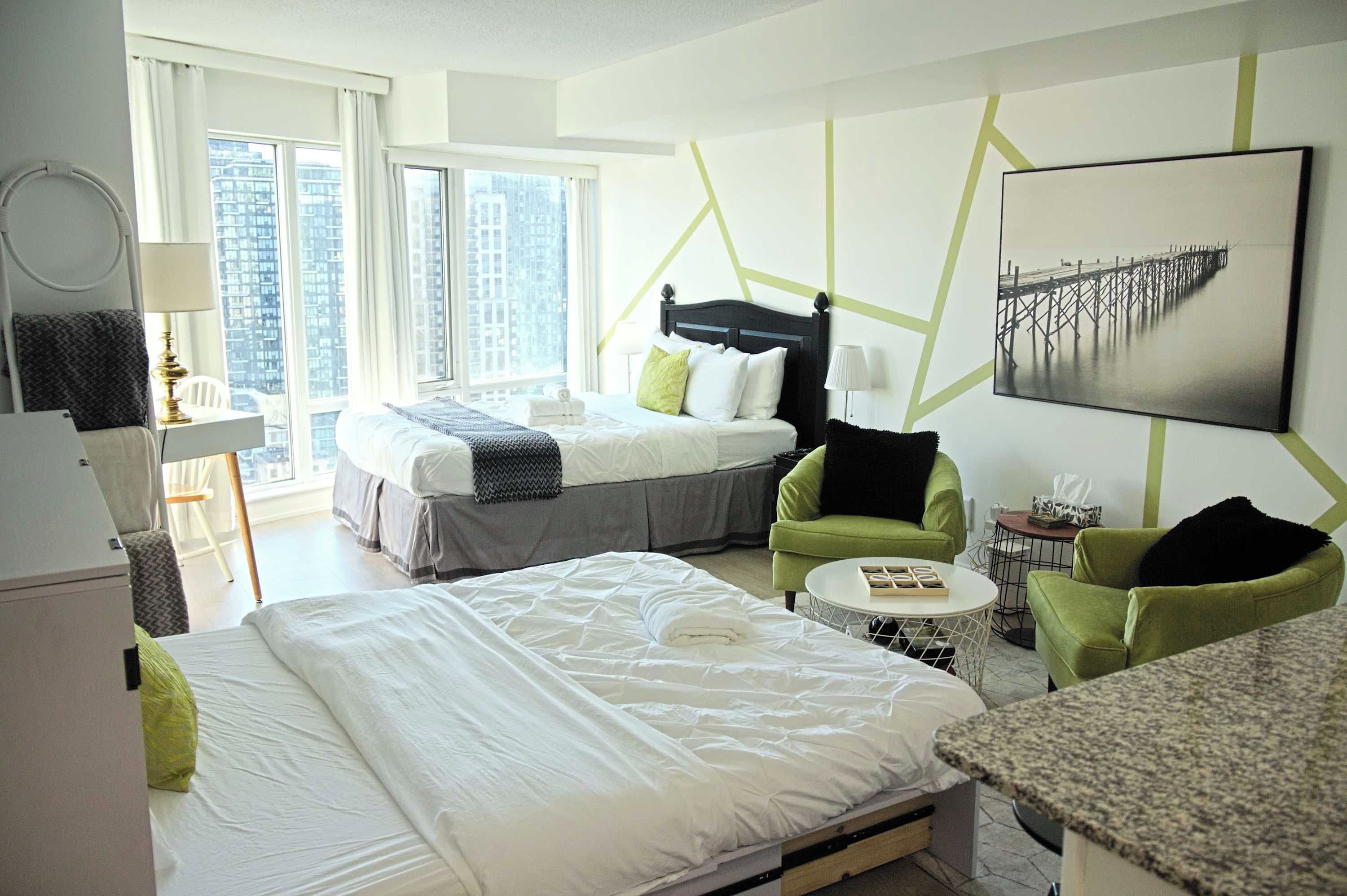 A hotel room with two beds, a desk, a chair, a small table, and a view of the city through large windows.