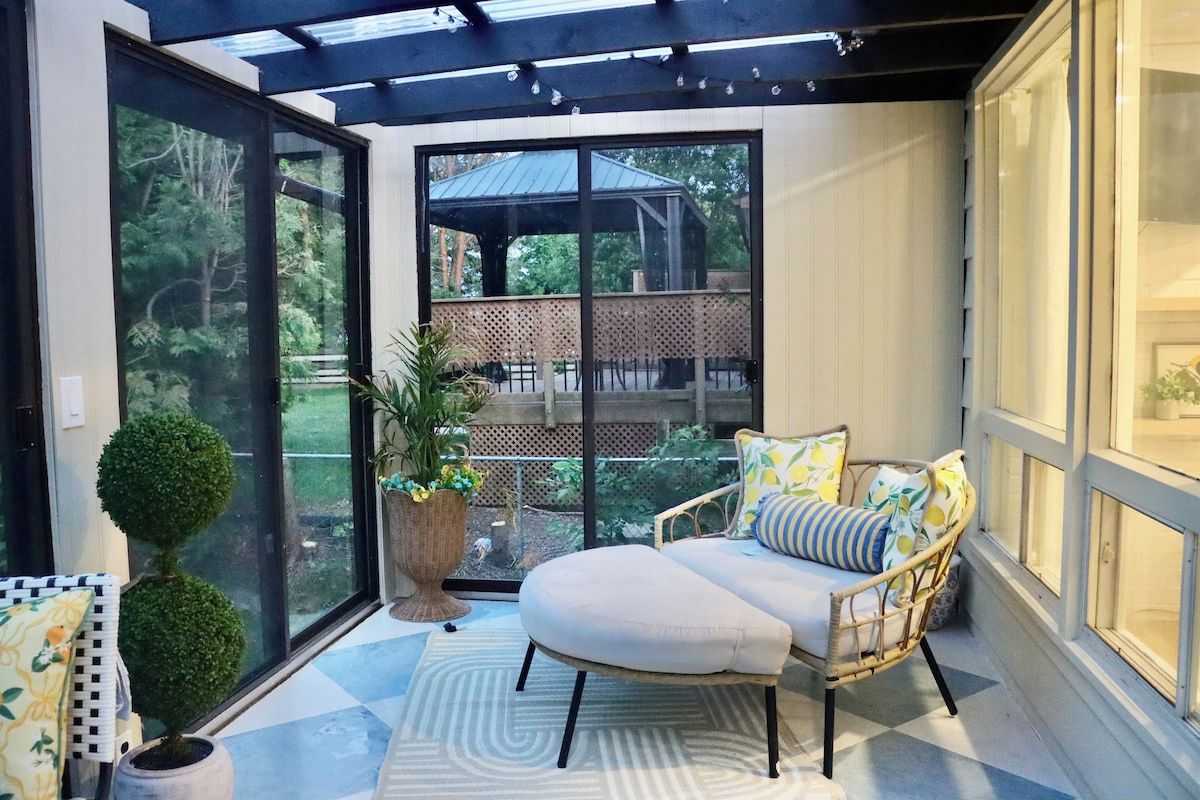 Eine sonnendurchflutete Terrasse mit Sitzgelegenheit mit Kissen, einem Blumentopf und Blick auf ein Holzdeck und Bäume.