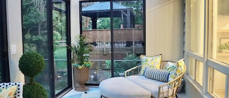 A sunlit patio with a cushioned seating area, a potted plant, and a view of a wooden deck and trees.