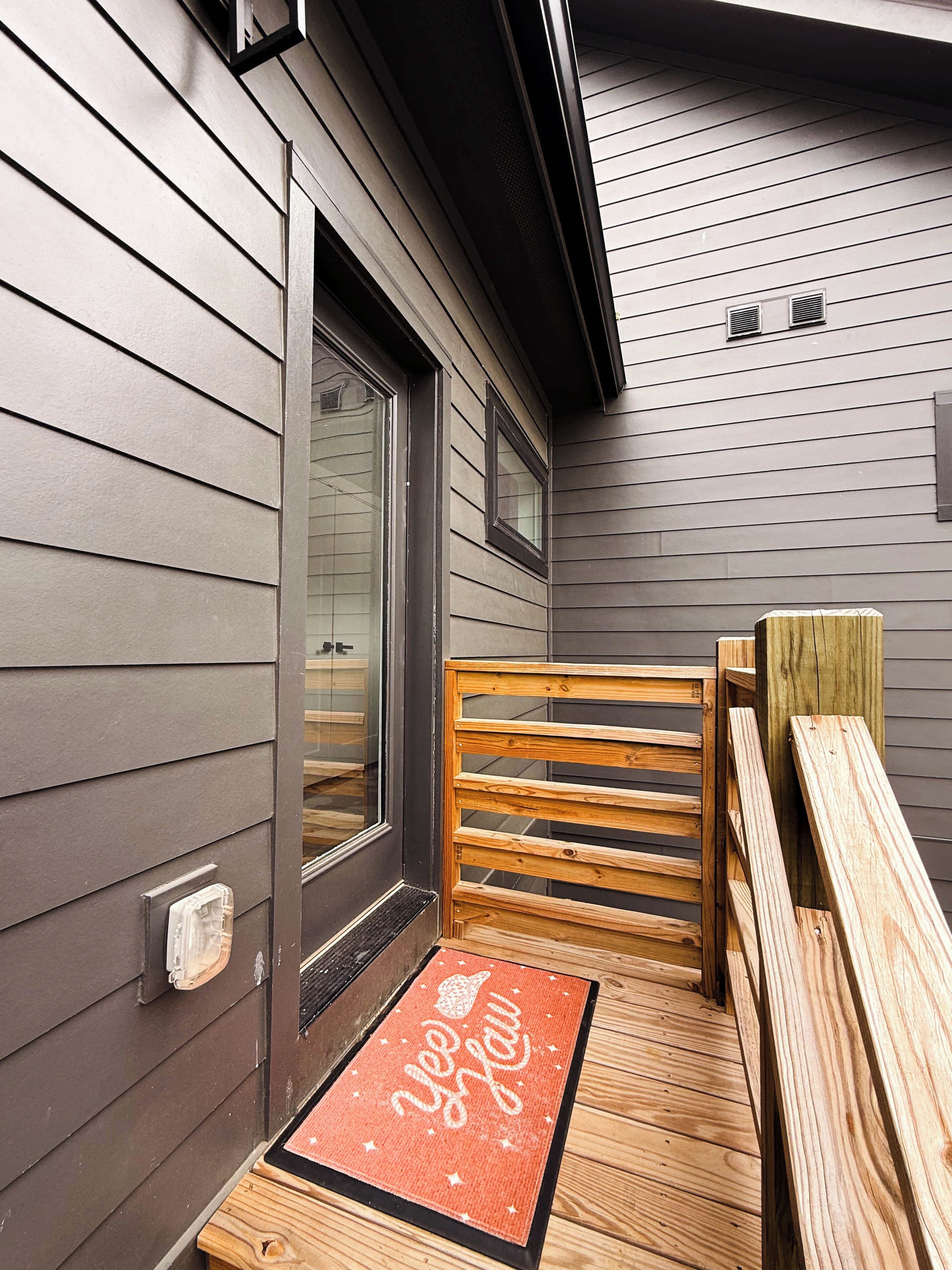 A cozy side porch with a wooden bench, warm lighting, and a vibrant red doormat, perfect for welcoming guests and enjoying quiet moments outdoors.