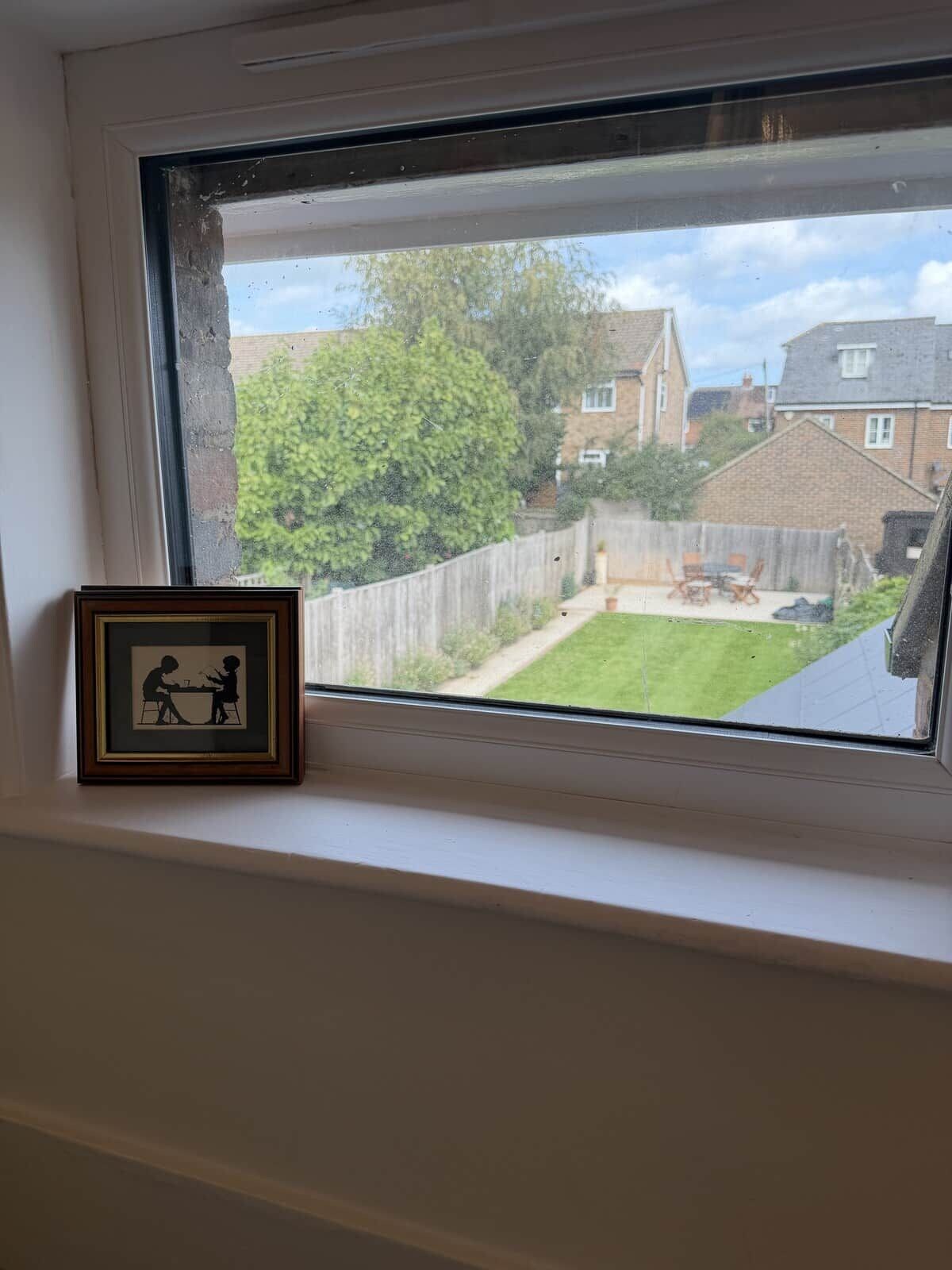 Een raam met uitzicht op een achtertuin, een houten hek en een lijst met een foto van twee personen.
