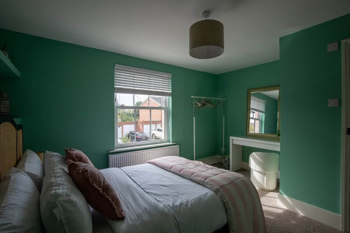 Ein Schlafzimmer mit einem Bett, einem Fenster, einer Heizung, einem Spiegel und einer weißen Mülltonne.