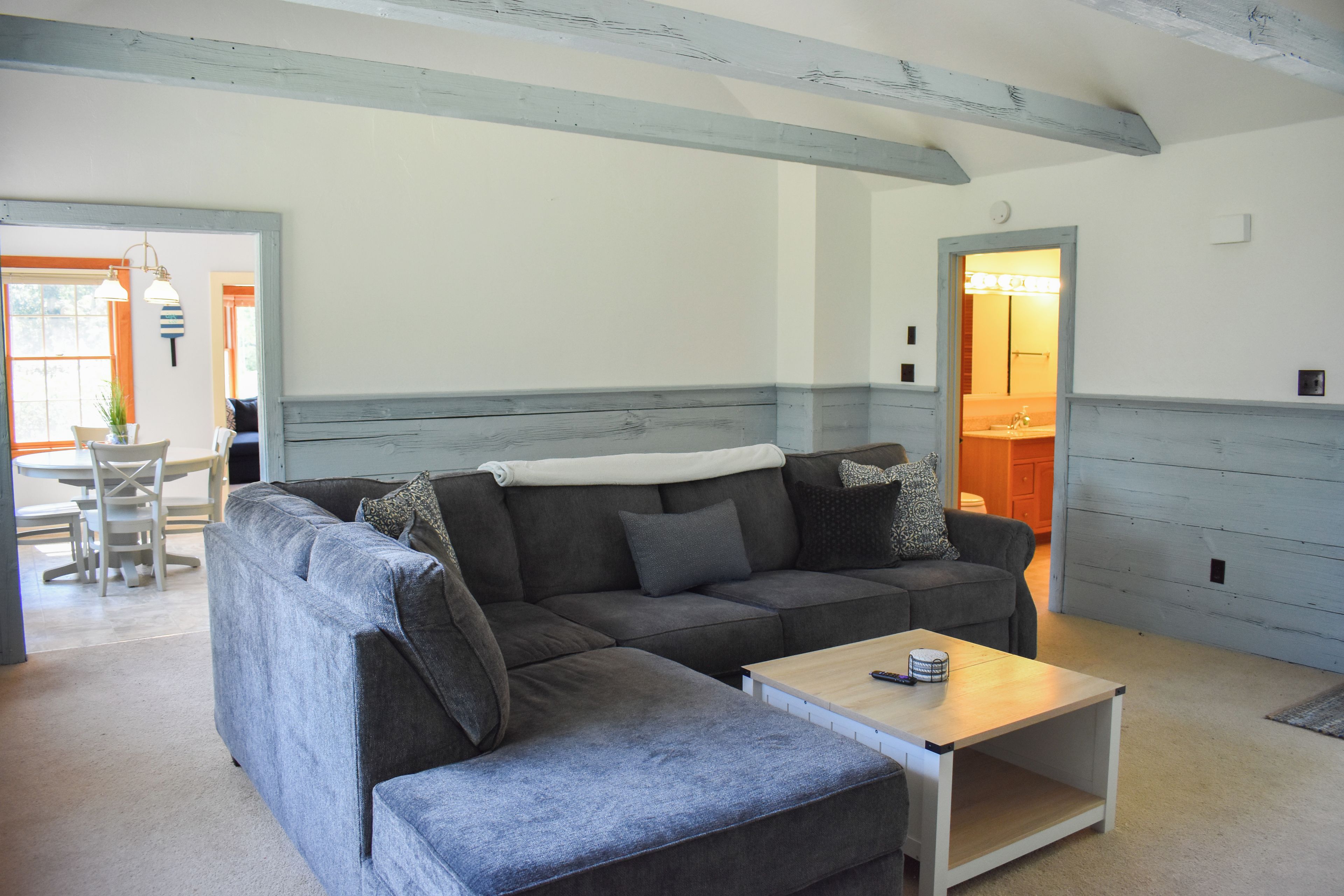 A living room with a grey sectional sofa, a small wooden coffee table, and a dining area with a white table and chairs.