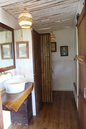Un baño con tocador de madera, un lavabo blanco y un espejo. Hay cuadros enmarcados en la pared y una puerta de bambú.