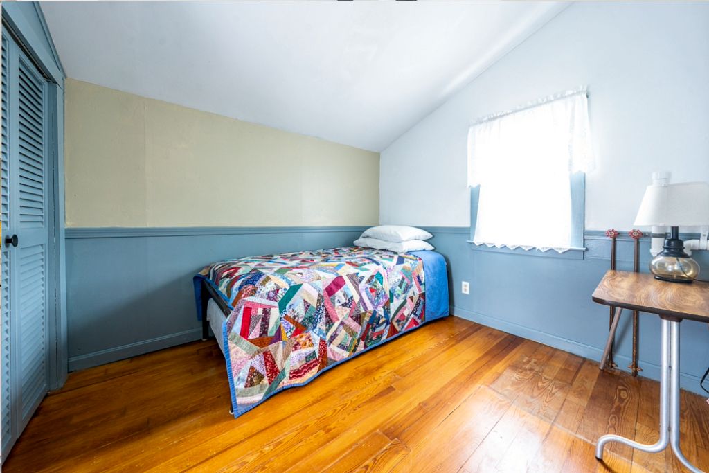 Une chambre avec un lit simple, une fenêtre avec des rideaux blancs, un plancher en bois et une petite table avec une lampe.