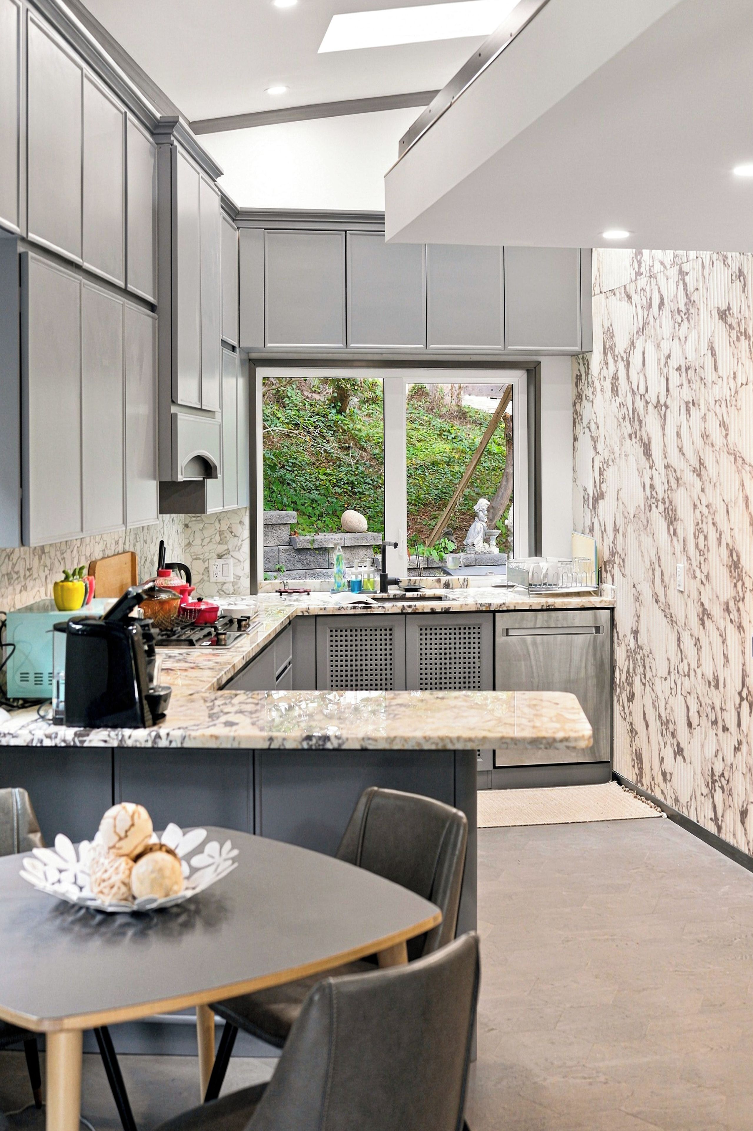A modern kitchen with grey cabinets, a marble countertop, and a window overlooking a garden.