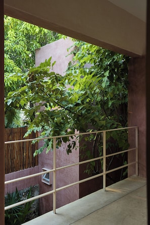 Un balcón con barandilla metálica, rodeado de exuberante follaje verde y una pared rosa.