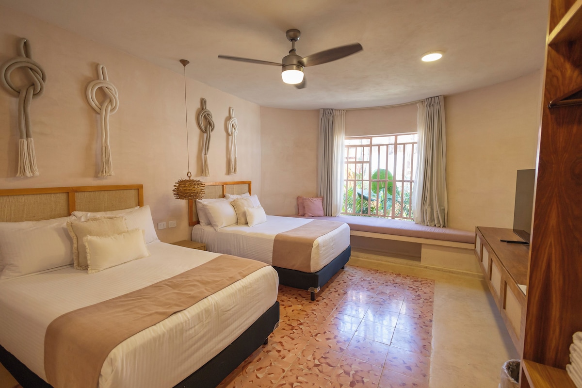Quarto de hotel com duas camas, ventilador de teto, janela com cortinas e televisão sobre um armário de madeira.
