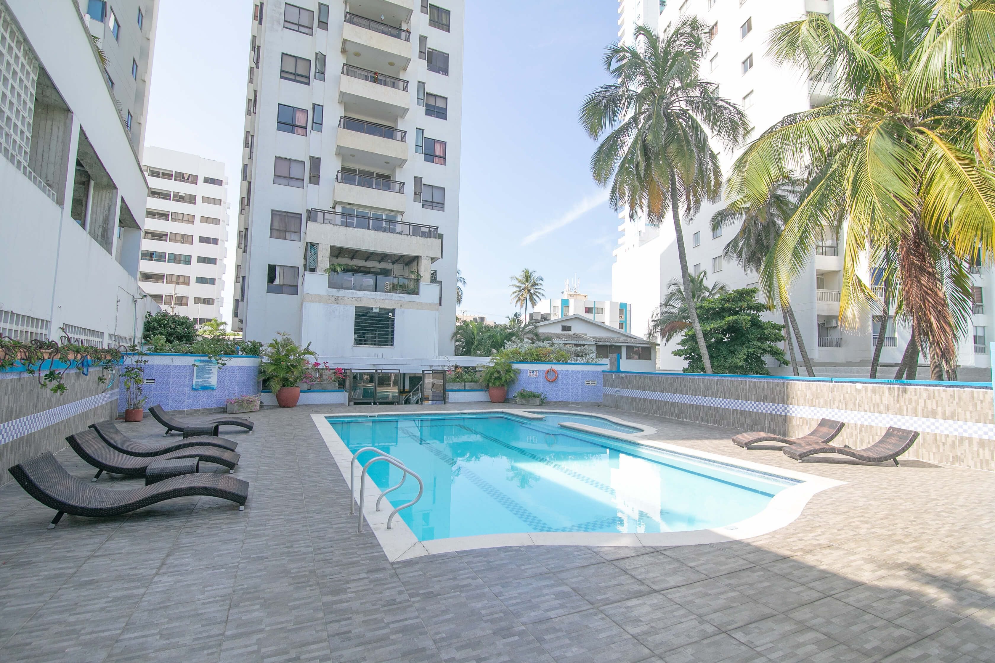 A swimming pool with lounge chairs, surrounded by a high-rise building and palm trees.