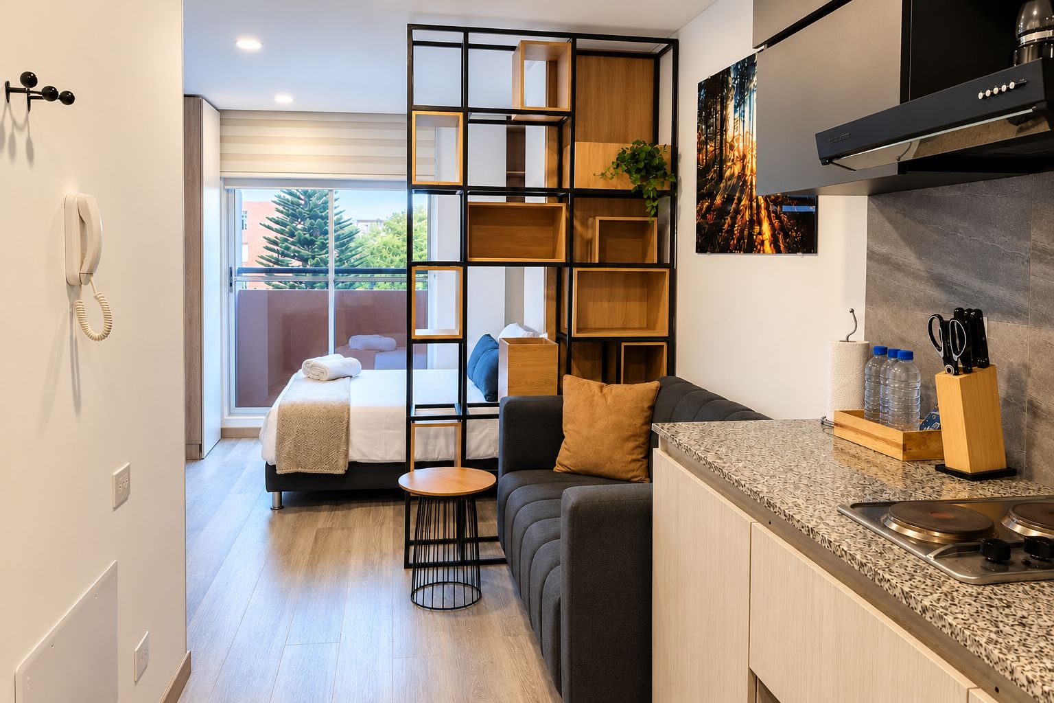 A modern hotel room with a kitchenette, a sofa, a dining table, and a large window with a view of greenery.