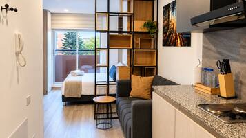 A modern hotel room with a kitchenette, a sofa, a dining table, and a large window with a view of greenery.