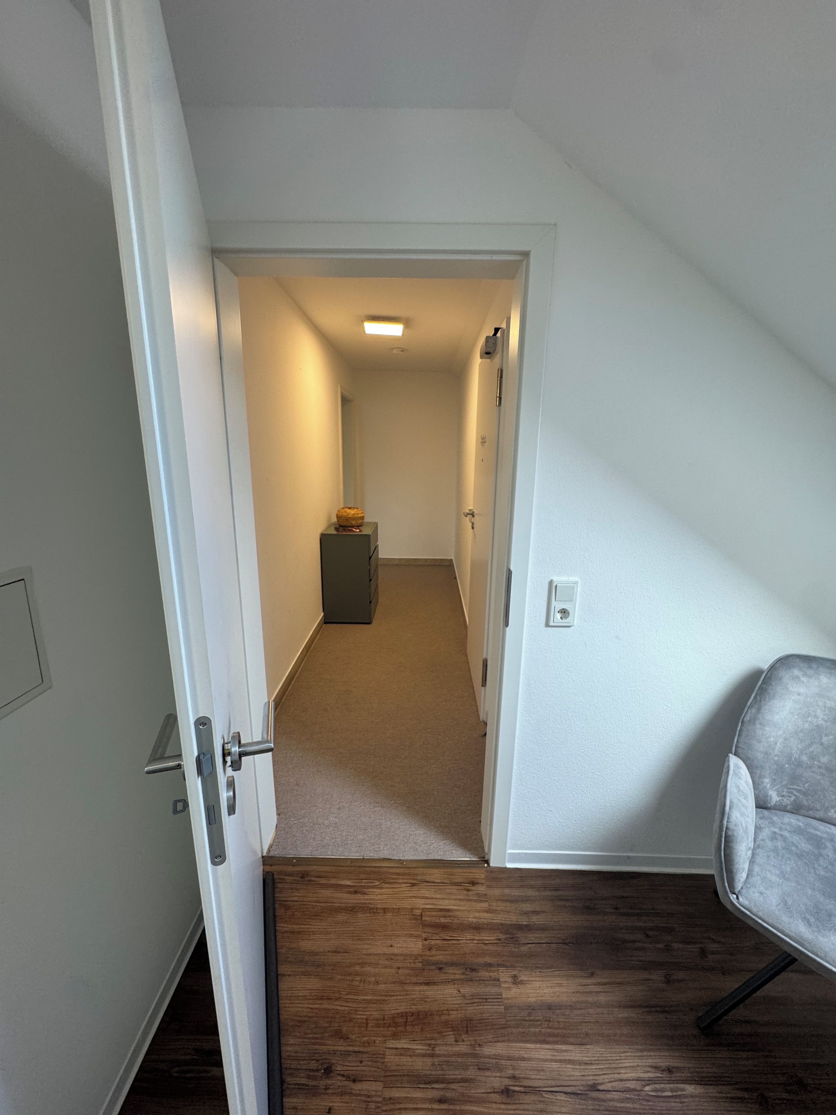 A narrow hallway with a wooden floor, a grey chair, and a door leading to another room.