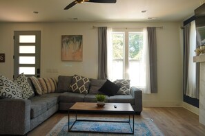 A living room with a grey sofa, a coffee table, a painting on the wall, and a window with curtains. - Pool and Games New Modern Home (Durham)