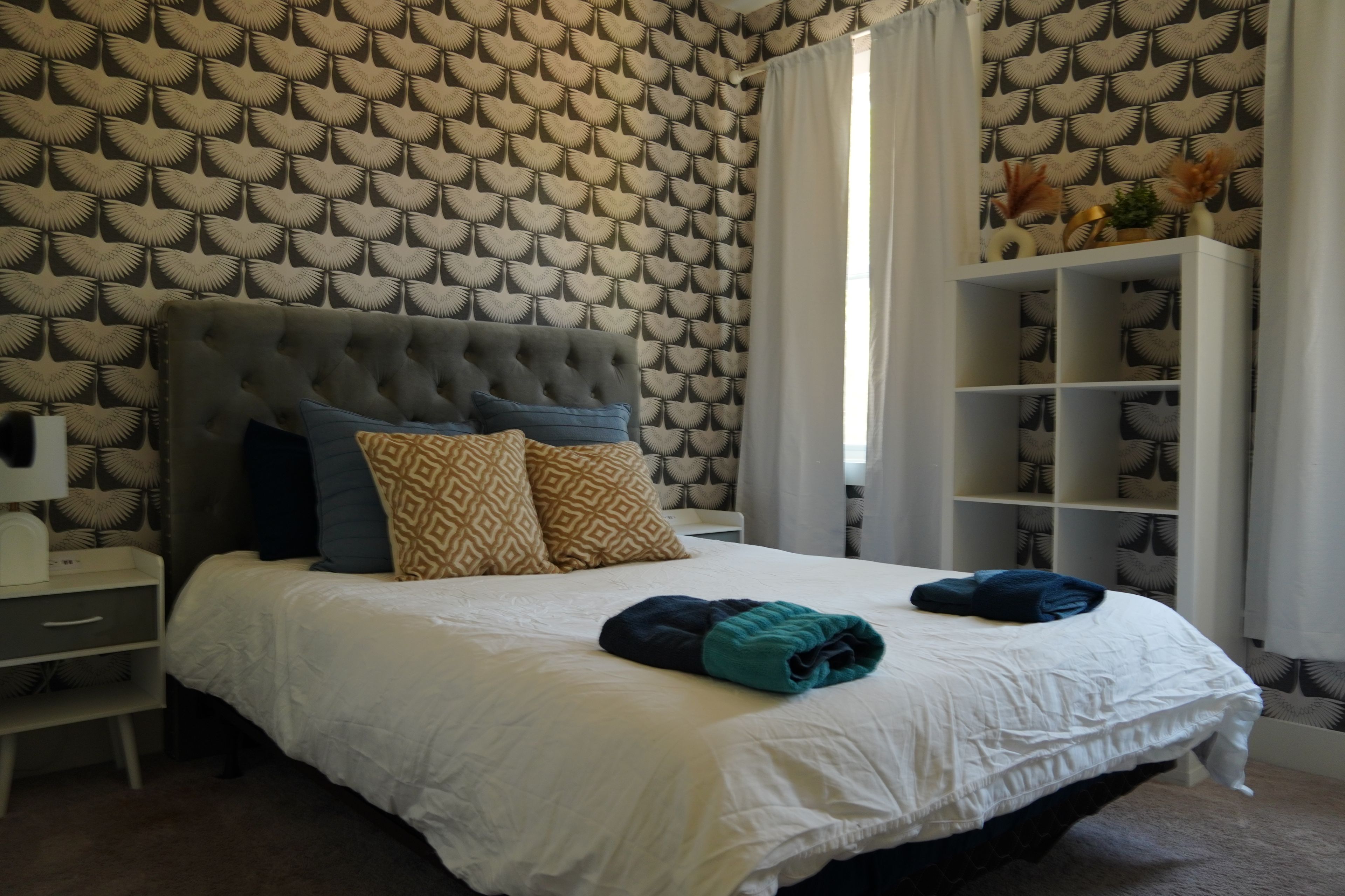 Un dormitorio con cabecera capitonada, papel tapiz estampado, cubrecama blanco y una mesita de noche con lámpara.