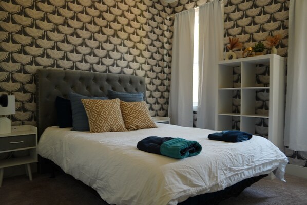 A bedroom with a tufted headboard, patterned wallpaper, a white bedspread, and a bedside table with a lamp. - Pool and Games New Modern Home (Durham)