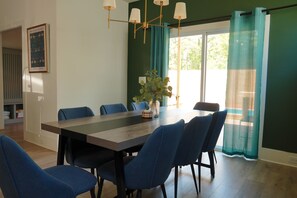 A dining room with a large table, blue chairs, a chandelier, and a view of the outdoors through a window with green curtains. - Pool and Games New Modern Home (Durham)