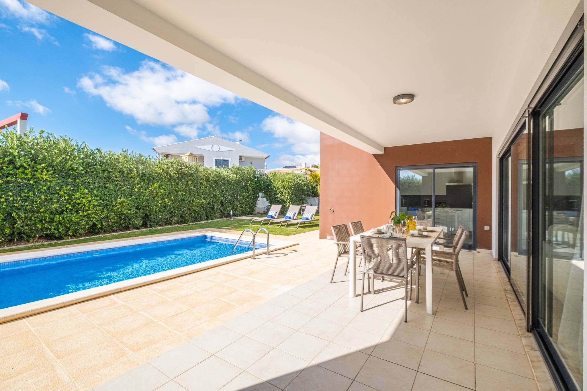 A modern outdoor pool area with a clear blue pool, a covered patio with a table and chairs, and a view of a white building in the background.