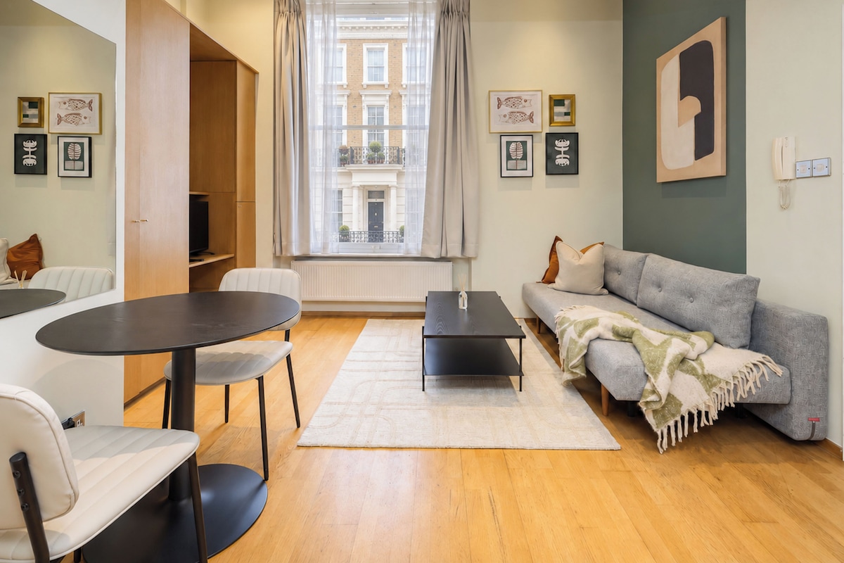 Ein modernes Wohnzimmer mit einer Couch, einem Esstisch und Stühlen. Ein Fenster mit Vorhängen, ein Heizkörper und gerahmte Bilder an der Wand sind ebenfalls vorhanden.