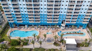Un hotel de varios pisos con un área de piscina, que incluye una piscina central grande y una piscina más pequeña contigua, rodeada de tumbonas y palmeras.