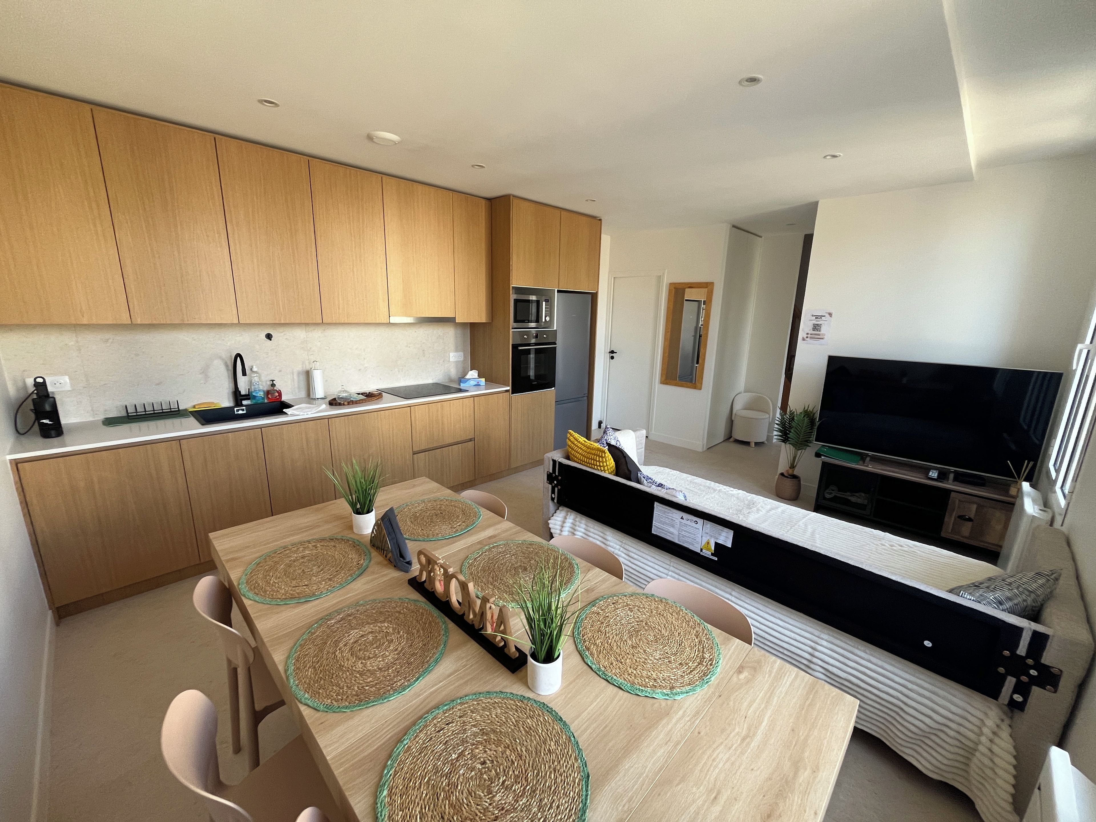 A modern kitchen with wooden cabinets, a dining area with a long table, and a living space with a TV and sofa.
