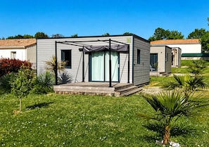 En moderne mobilhome med en overdækket terrasse, omgivet af en velplejet græsplæne og små træer.