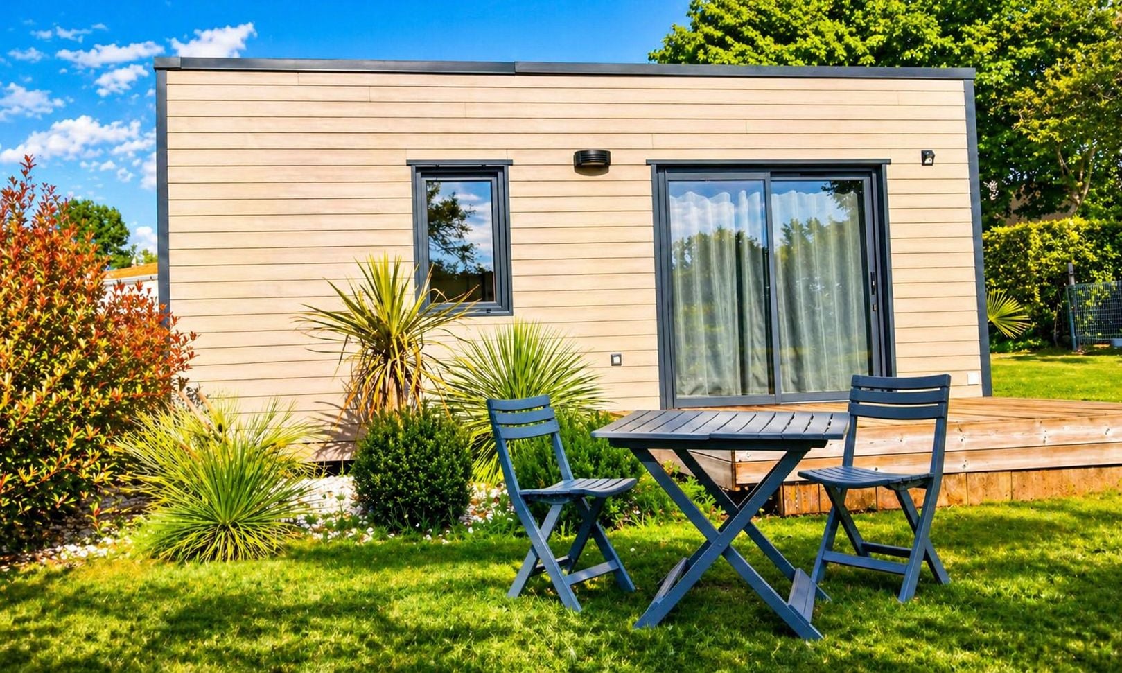 Un bâtiment moderne, de plain-pied, doté d’un toit plat, d’un petit patio avec une table et des chaises, et d’une fenêtre avec un rideau.