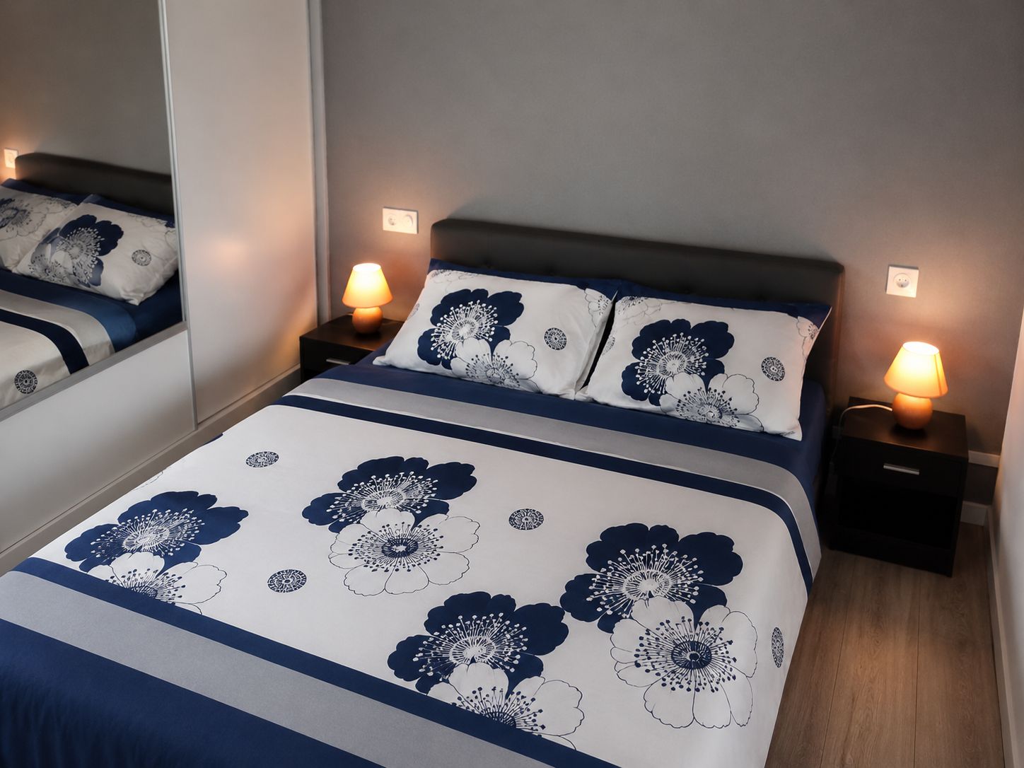 Un lit bien fait, avec un couvre-lit à motifs floraux, deux lampes de chevet et un miroir au mur.