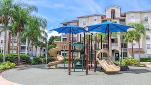 Un parque infantil con toboganes y estructuras para escalar, rodeado de palmeras y un edificio con balcones.