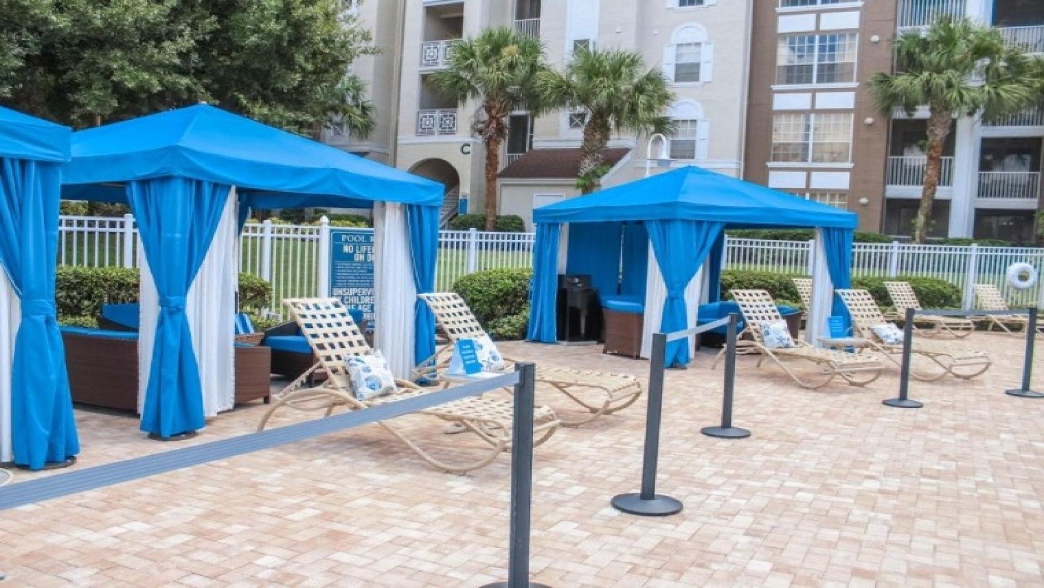 Un área junto a la piscina con toldos azules, sillones de descanso y suelo de baldosas.