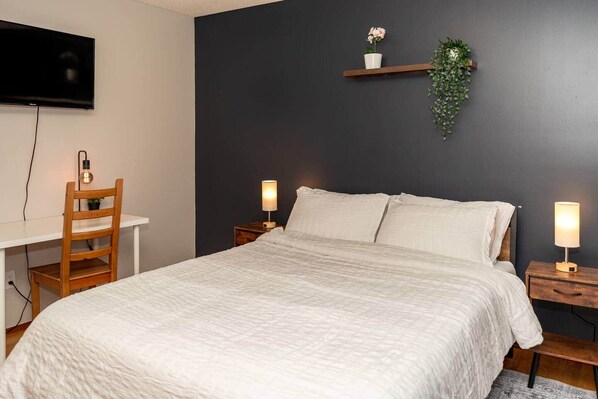 Une chambre à coucher comprenant un lit, des tables de chevet, un bureau, une chaise, un téléviseur et une étagère murale agrémentée d’une plante.
