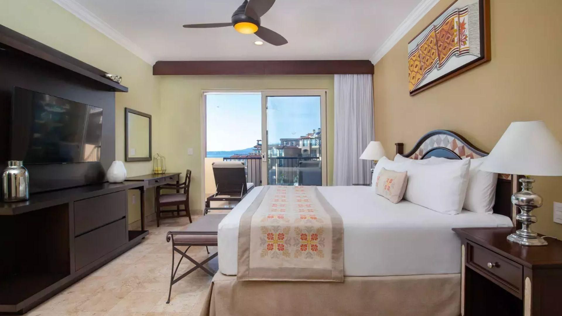 Habitación de hotel con cama, escritorio, silla, televisor y balcón con vistas.