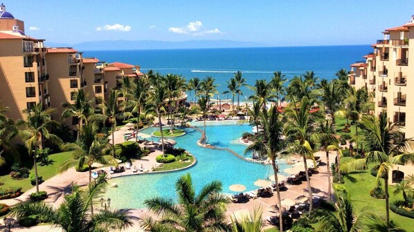 Um resort com piscina central, palmeiras e vista para o oceano.