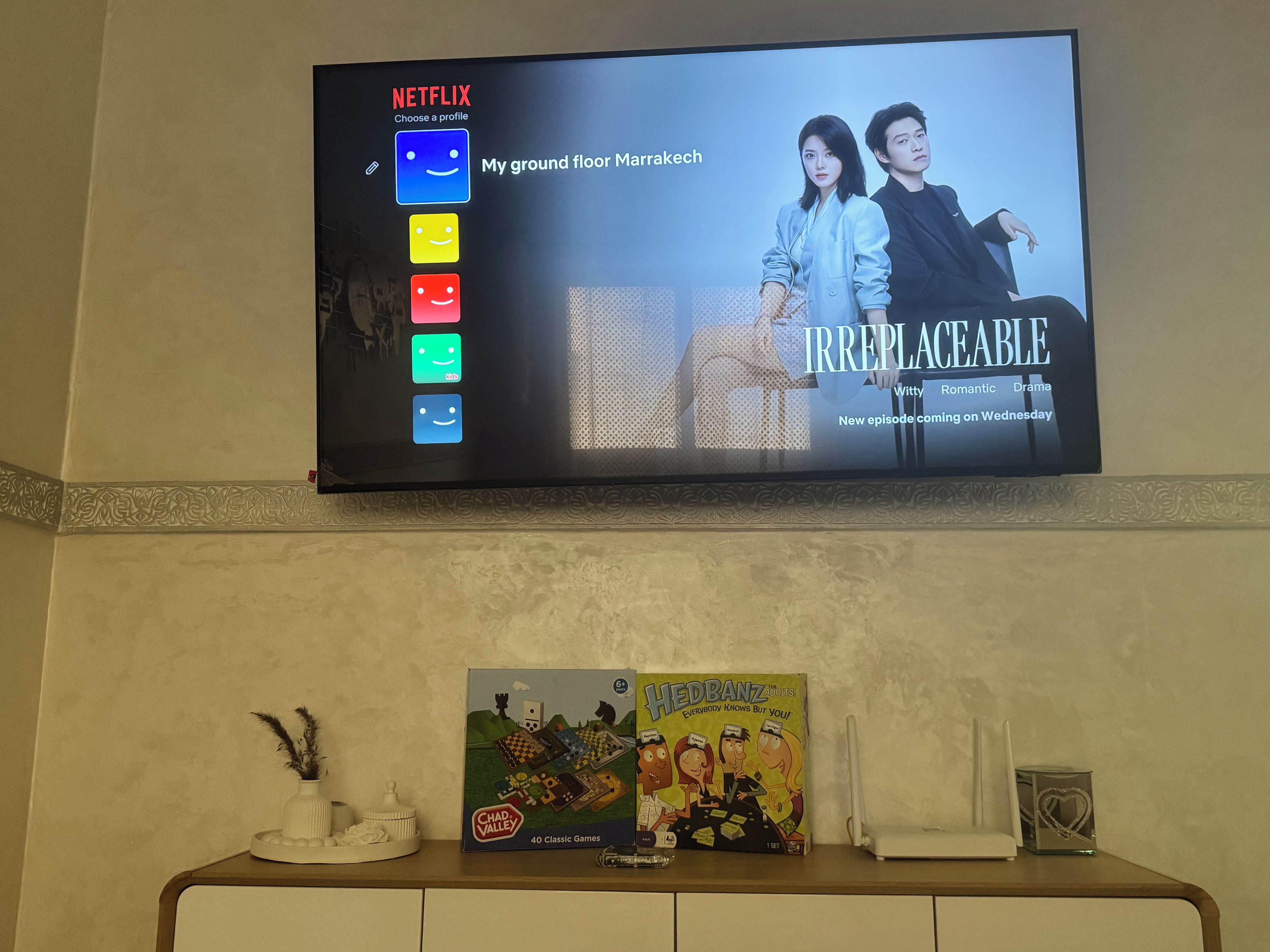 A television displaying a Netflix profile with the title 'My ground floor Marrakech' and the show 'Irreplaceable' with a subtitle 'Witty, Romantic, Drama'. Below the TV, there is a table with a board game titled 'Hedbanz' and a decorative vase.