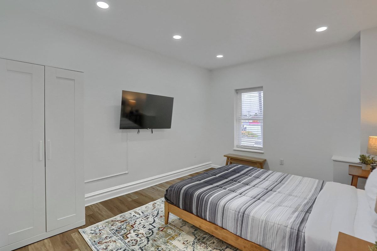 Een slaapkamer met een bed, een aan de muur bevestigde televisie, een raam met zonwering en een klein nachtkastje met een lamp.