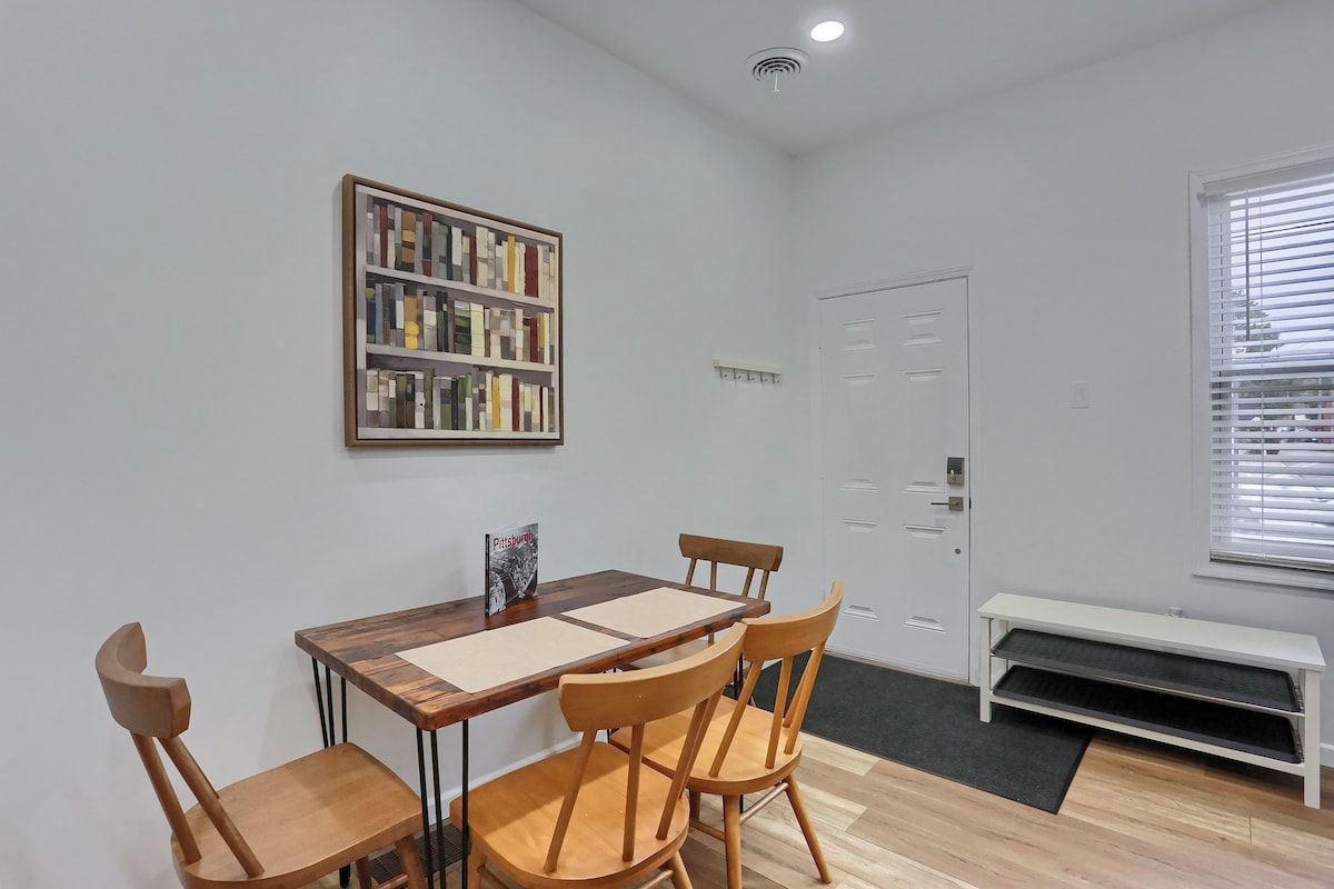 Een eetgedeelte met een houten tafel en stoelen, een boekenkast, een deur en een bankje.