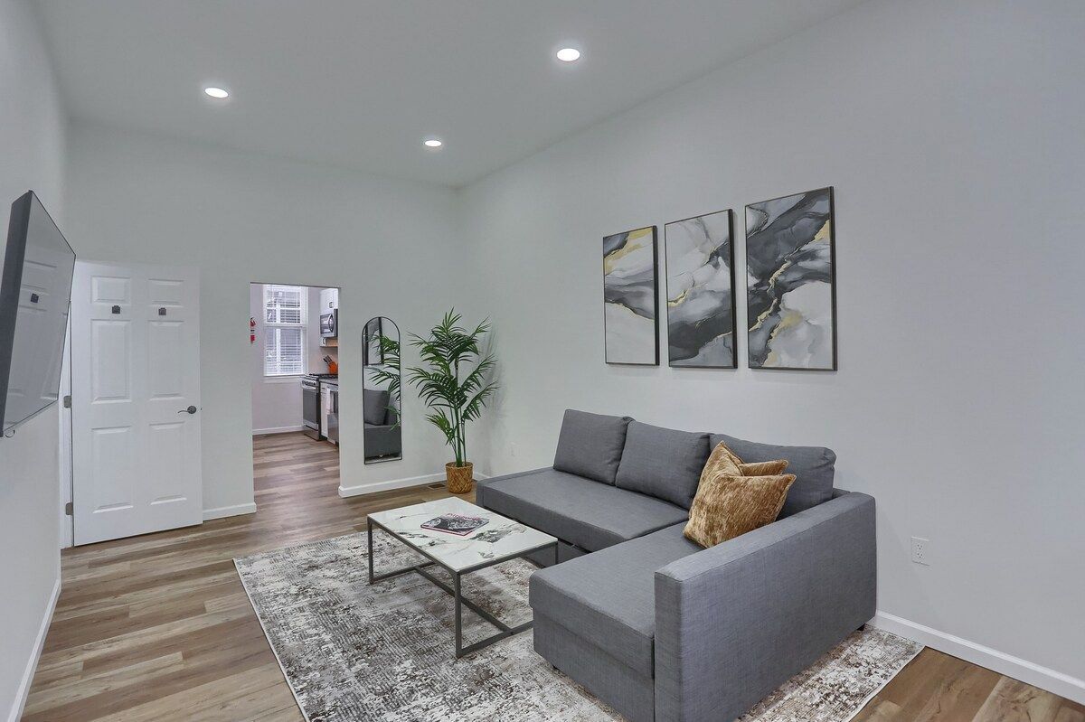Een moderne woonkamer met een grijze bank, een salontafel en een grote spiegel. Aan de muur hangen abstracte schilderijen en er staat een potplant.