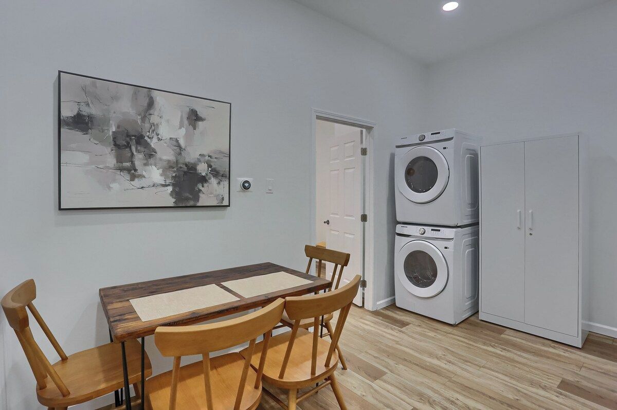 A room with a dining table, chairs, a wall-mounted abstract painting, a washing machine, and a wardrobe.